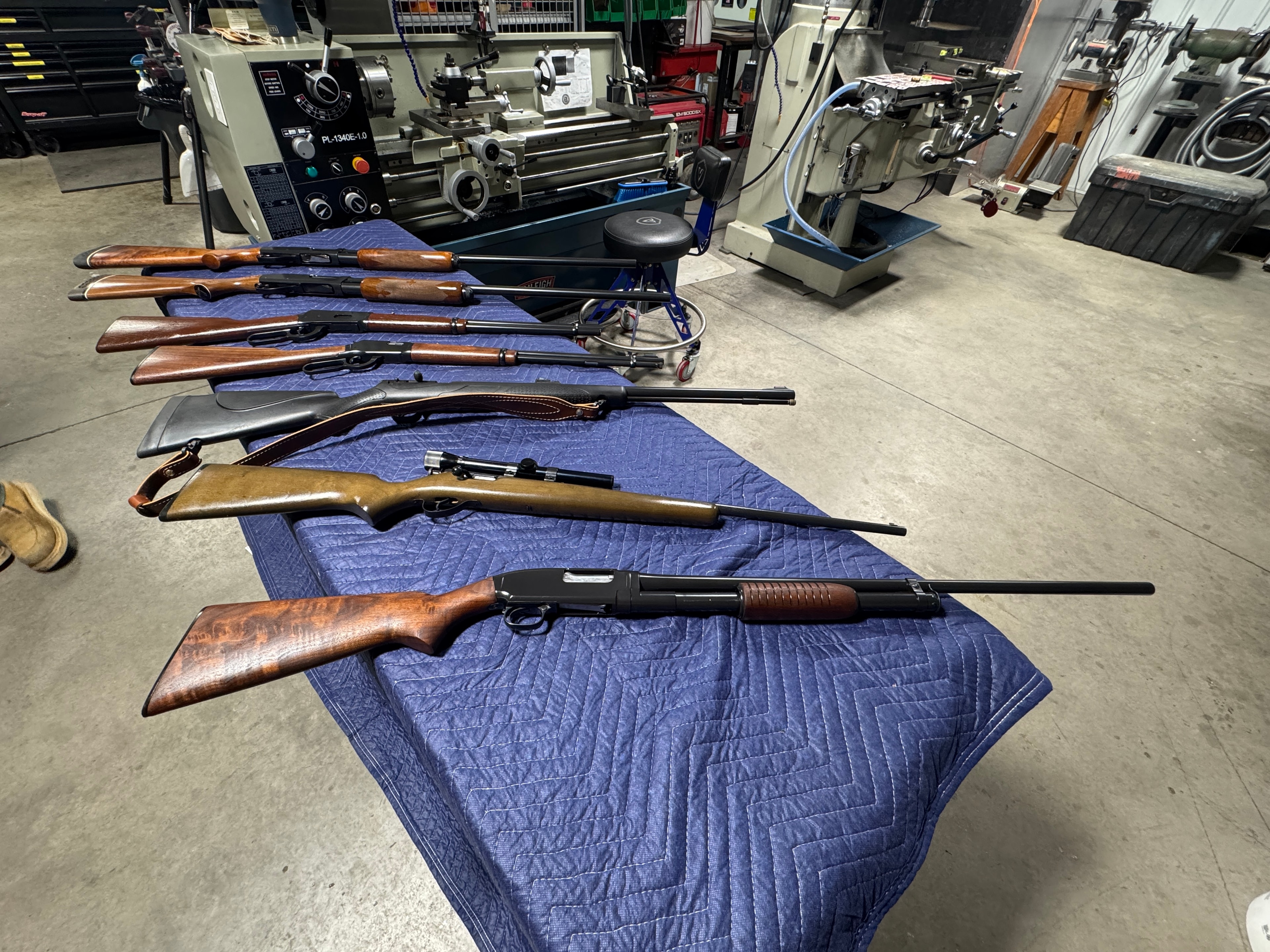 Estate collection of rifles and shotguns laid out on a blue mat in the Northwoods Firearms machine shop — Baileigh lathe in background